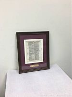 Framed bible page on white table against a plain wall, showing overall lot presentation.