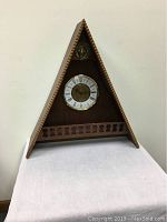 Front view of the triangular wooden mantle clock displaying clock face, metal lion head, and decorative wooden trim.