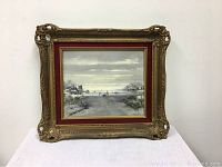 Frontal view of the oil on board painting depicting a Dutch winter skating scene with windmills, housed in ornate gold gilded frame sitting on table.