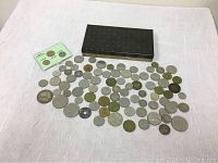 Photo showing the spread of approximately 70 vintage foreign coins laid out with a rectangular tin and a small display card with two coins.