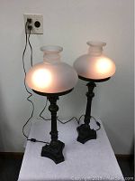 Pair of mid century lamps with smoked glass shades lit, on white surface, showing frosted bulbous shape and dark metal bases with cords and inline switches.