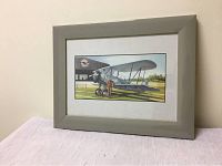 Framed watercolor artwork showing the full piece with biplane and pilot, displayed on white surface against wall.