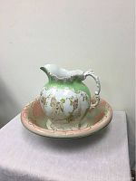 Pitcher and bowl set displayed on a cloth-covered surface with a light gray background; visible ornate painted details and color.