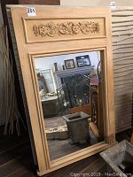 Full view of the large wood framed mirror leaning against a wall among other furniture and decor, showing the raised carved wood panel on top and the full mirror glass below.