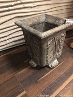 Front angled view showing square concrete planter with ornate fluted sides, claw-footed base, and antiqued finish.