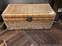 Front view of rectangular wicker chest showing woven wicker design and brass-like latch with wear and holes visible.
