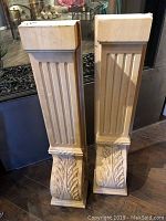 Two wooden shelf bracers standing side by side, showing front fluted and leaf carvings.