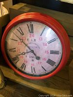 Full view of round reproduction clock with red metal frame under glass, Roman numeral and 24-hour dial face