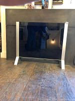 Shows front view of freestanding black smoked glass firescreen with silver metal framing and feet standing on wood floor.