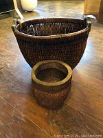 Two woven baskets of different sizes, dark brown, placed on wooden floor. Larger basket with handles and smaller basket with solid rim.