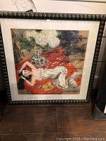 Photo showing framed watercolor artwork of a woman reclining on red throw under a vase of flowers.