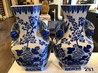 Pair of blue and white ceramic vases with detailed floral, fruit motifs and sculpted leaf handles on a white background, displayed on a white surface with wooden flooring beneath.
