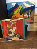 Two artworks shown: a colorful Picasso print in a gold frame in front, and a vibrant acrylic landscape painting on canvas behind.