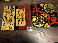 Two oriental plaques with calligraphy and symbols displayed on floor. One has black calligraphy on gold and red background. Other has red calligraphy and symbols in gold circles on black background.