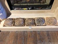 Five wooden square plaques placed in a row in front of a fireplace, showing the carved floral and round mound designs and overall size.