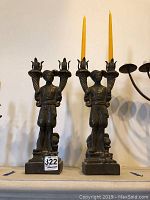 Pair of resin candleholders resembling men holding cornucopias, with tall yellow candles placed in each holder.