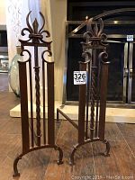 Pair of brown metal andirons with fleur-de-lis decorative tops in front of a fireplace.