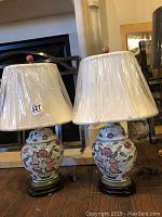 Pair of ceramic table lamps with cream cloth shades still wrapped in plastic, showing floral design and wood bases. One lamp base shows damage.