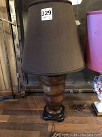 Full view of the lamp with dark cloth shade, sitting on wooden floor beside a pink stool, showing the whole lamp and electrical cord.
