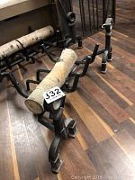 Black wrought iron log holder on wooden floor with birch logs placed inside to demonstrate use. Shows sturdy legs and design details.