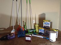 Full lot photo showing the cleaning tools lined up: Shark handheld vacuum, broom and dustpan, mops, Swiffer boxes, spinning scrubber system components, garbage can.