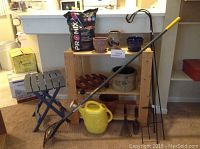 Photo showing a gray folding table, yellow watering can, garden tools including hand rake, spade, and a long-handled narrow rake, bag of potting soil, multiple ceramic pots, large crock, clay pot, wooden plant caddy, and metal shepherd's hooks.