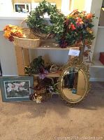 Various silk florals including greenery wreath, silk flower bunches, wicker baskets, gold tone oval mirror, and framed floral print arranged on and in front of wooden shelves