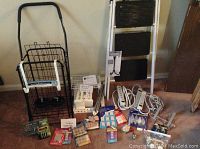 Overall view of various household hardware items including grocery cart, step ladder, surge protectors, hooks, batteries, and candles arranged on floor and leaning against wall.