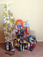 Full view of ironing board, two irons, metal shelf loaded with cleaning supplies, bucket with supplies on floor