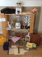 Overall view of wooden shelving holding multiple kitchen appliances and items including Oster hand mixer, blender, Cuisinart chopper in box, baking dishes, towels, graters, cutting board, muffin pan.