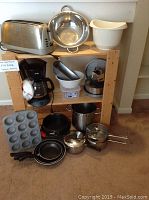 Full view of kitchenware lot on wooden shelving unit including toaster, coffeemaker, pots, pans, colander, muffin tin, mixing bowls, grater, and baking items.