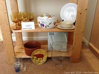 Shelf with six amber wine glasses, Royal Worcester Dutch oven with fruit design, Lenox bowl and platter, brown ovenware, shot glass set, and glass trifle bowl.