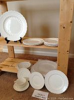 Group of dishes on wooden shelf including stacked dinner plates, bread/butter plates, saucers, one cup on saucer, and salad plates laid out in front.