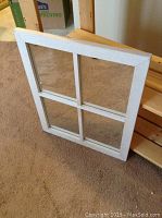 Square white wooden frame with four glass panes separated by muntins shown leaning against shelving and carpeted floor.