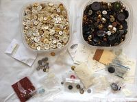 Photo showing two containers filled with many white, cream, and dark buttons, with additional small loose bags of buttons and some packaged buttons.