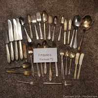 Full view of the flatware assortment laid out on carpet including knives, forks, spoons, butter knives, and serving spoons.
