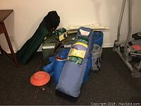 Photo of camping gear arranged on carpeted floor including twin size portable bed in blue case, two sleeping bags, two lawn chairs, cooler and lantern.