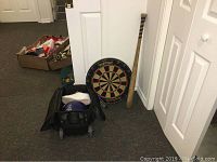 Overall view showing dartboard with darts, wooden baseball bat leaning against wall, black rolling bag with purple bowling ball inside, and white woman's shoes on the floor.