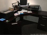 Corner desk set up with computer monitor, printer, keyboard, and other office accessories showing the L-shaped design and riser area.