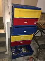 Blue plastic drawer units with red, yellow, and blue drawers stacked. Bottom drawer open showing loose hardware and tools inside.