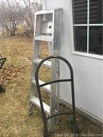 Side view of the aluminum four step ladder leaning against an exterior wall next to the black metal dolly.