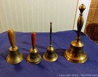 All four vintage brass bells displayed upright on a blue cloth surface, showing the variety in sizes and handle shapes.
