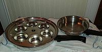 View of Canadiana surgical steel deep pan with egg poacher insert and a medium frying pan on a fabric surface.