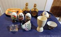 Photo showing two hand painted eggs on black stands, three ceramic eggs, two gold-tinted glass decanters with animal figurines, peach-colored tray, clear glass paperweight, ceramic fish, white egg cup, and small floral bowl.