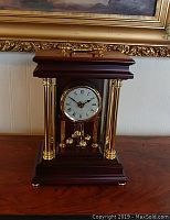 Front view of quartz mantle clock with roman numeral dial, gold columns, and motion pendulum visible through glass sides.