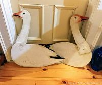 Pair of white plywood snow geese silhouettes leaning against a door, showing front side with painted orange beaks and black detailing.