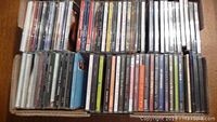 Photo showing three rows of CD cases in a box, viewed from above, showcasing multiple CD spines with mixed artist names visible.