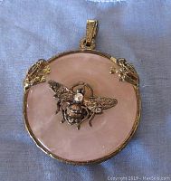 Top side of round pink glass pendant showing two metal frogs on the edges and a metal bee with rhinestone in the center in silver-tone setting.