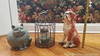 Three decorative statuettes: a gray ceramic cat, a reddish-brown and white ceramic dog, and a metal birdcage candle holder with a candle inside. A measuring tape stands behind the dog statuette for scale.
