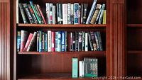 Photo of a wooden bookshelf containing various hardcover and paperback books arranged in two rows with some books standing vertically on the bottom shelf.